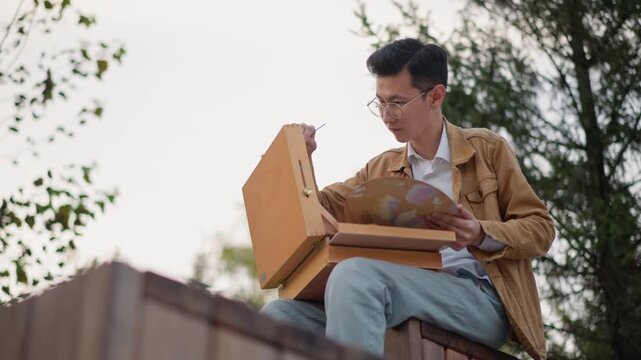 asian painter painting on rooftop captured in golden light, street art session with wooden box easel and round oil palette, artist seated on ledge mixing colors with focused hands, casual jacket,