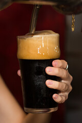Close-up of dark beer being poured into a glass with thick foam. Hand holding glass during pouring process, fresh cold alcoholic beverage, bar drink concept, selective focus.