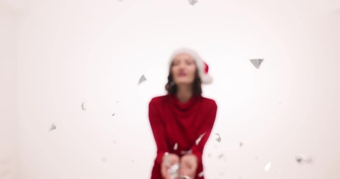 Woman in Santa hat holds handful of confetti near face. Holiday model blows paper pieces forward creating sparkling explosion around head slow motion