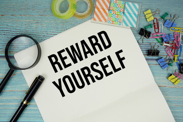 Reward Yourself text on a paper surrounded by office supplies and magnifying glass on a blue wooden...