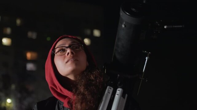 urban night stargazer with telescope on rooftop, female wearing red hood and glasses studies sky through large refractor, city lights and bokeh background, contemplative expression, slow pans