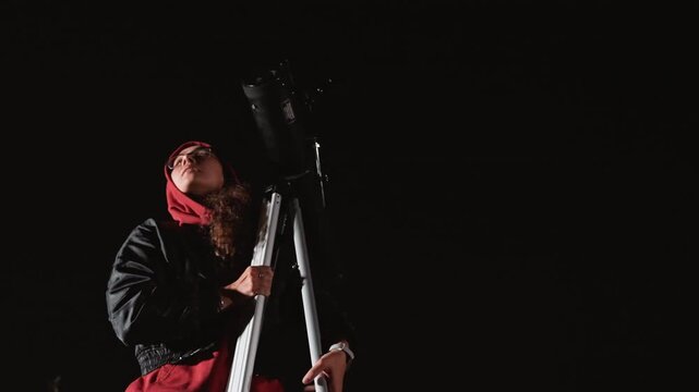 night sky stargazer with telescope and tripod adjusts eyepiece in grassy urban field beside lit dormitory windows, hooded observer focuses on stars and constellations, quiet atmosphere of curiosity