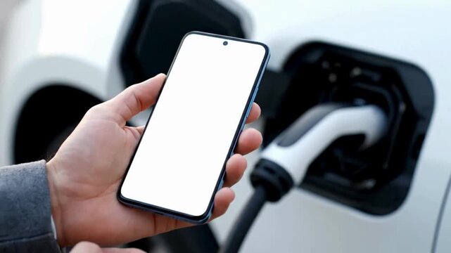 Modern electric vehicle charging while a user holds a smartphone with a blank screen, symbolizing the seamless integration of smart technology into sustainable automotive energy solutions