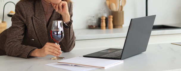 Woman at the table with a glass of wine in her hands, laptop on the table. Business woman, online meeting. Work from home, online. Unrecognizable woman in a suit with a glass of red wine