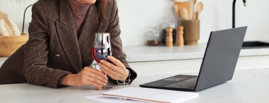 Woman at the table with a glass of wine in her hands, laptop on the table. Business woman, online meeting. Work from home, online. Unrecognizable woman in a suit with a glass of red wine