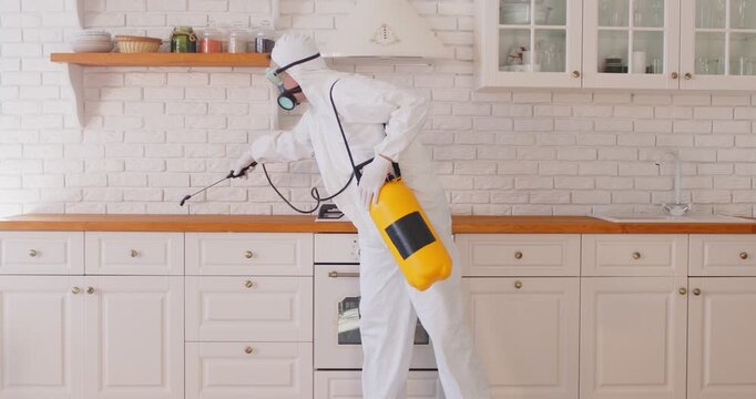 Professional exterminator in white protective suit and mask uses sprayer to treat stove and kitchen cabinets with pesticide for insect control. Pest control services concept