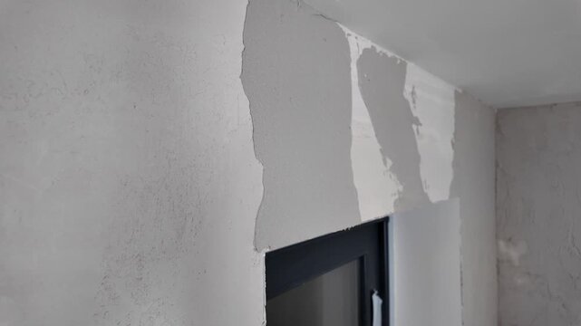 A person is repairing a wall by applying plaster to cover damaged areas. This takes place indoors, near a window, during daylight hours.