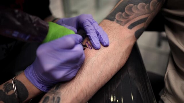 Professional tattoo artist wearing purple nitrile gloves meticulously inks the name belen on a man's forearm, using a tattoo machine with black ink in a modern and clean tattoo parlor