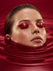 Surreal beauty portrait of a woman with glossy red eye makeup, partially submerged in red liquid for a calm emotional concept