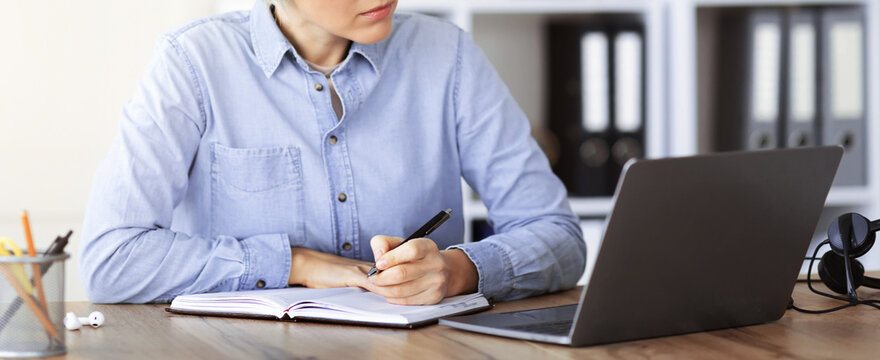 A woman sits at a wooden desk, writing in a notebook while looking at a laptop screen. She has headphones nearby and is focused on her task in an office space with filing cabinets.