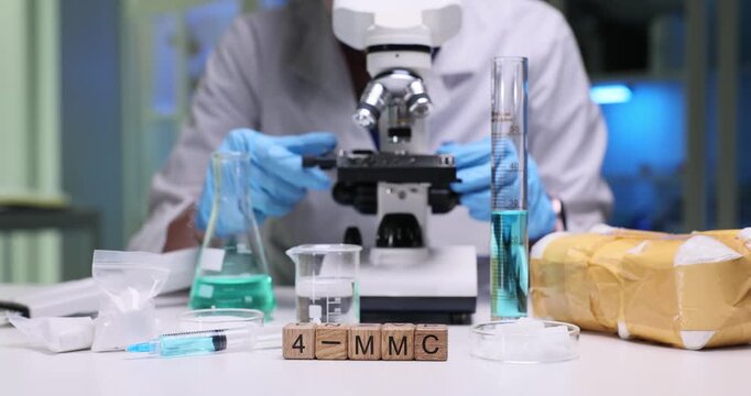 Wooden cubes spell code 4 MMC near colorful lab glassware. Scientist adjusts microscope near pack of drugs by test tubes and liquid samples in lab