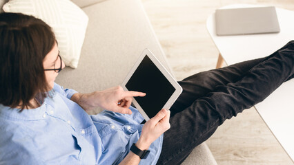 A person sits on a sofa with their legs stretched out. They are focused on a tablet held in one...