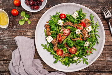 Warm beef meat salad with arugula, lettuce, feta and tomatoes on a plate on wooden background, top view. A delicious gourmet dinner in the rustic style. High quality photo