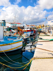 Fishing boats moored in Marsaxlokk harbor, with houses, church, blue sky and white clouds in the background