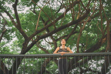 Asian fitness woman closing eyes under green trees. Mindfulness outdoor workout relaxation and mental wellness concept. Healthy female athlete enjoying fresh air in a park