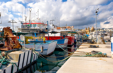 Fishing boats moored in Marsaxlokk harbor, with houses, church, blue sky and white clouds in the background