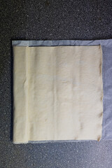 A rolled sheet of puff pastry sits on a gray speckled surface, partially on baking paper.