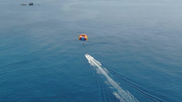 Aerial top view of speed boat towing colorful parachute with person parasailing over deep blue open sea water on sunny summer day