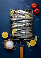 Fresh sardines arranged in a silver grilling basket on a dark blue background with lemons, salt, tomatoes, and peppercorns in a flat lay. Generative AI