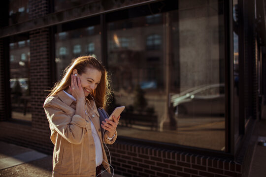 Smiling young woman listening to music on smartphone on city sidewalk