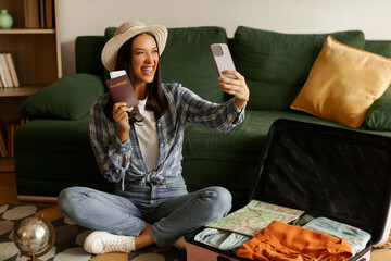 Smiling woman taking selfie with passport on cellphone while sitting near open suitcase, preparing...