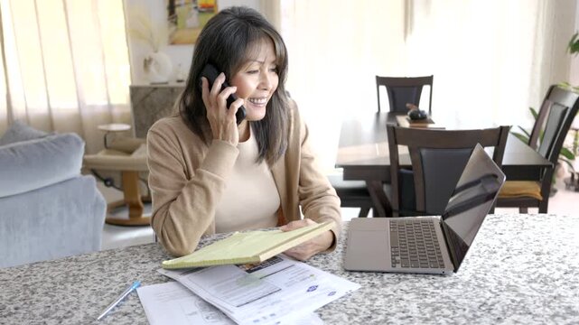 Attractive mature woman holding note pad enjoying talking business on cellphone sitting at home with conversation on finance in technology