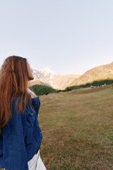 Obraz premium Woman redhead mountains meadow denim outdoors, profile portrait of young woman standing in grassy field, looking toward distant peaks at sunrise for travel, hiking and peaceful nature escape.