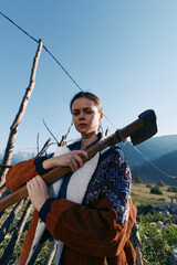 Fototapeta premium Woman with axe outdoors in rural mountains, portrait by wooden fence in a village setting, lumber tool and nature scene with rustic clothing and determined gaze.