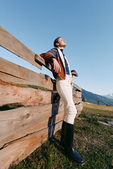 Obraz premium Woman leaning on wooden fence in countryside with mountains and clear sky, wearing knit sweater and rubber boots. Relaxed outdoor portrait, nature view and pastoral fashion.