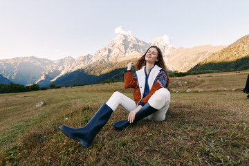Obraz premium Woman sitting on mountain meadow smiling in colorful jacket and rubber boots, relaxed travel portrait in nature with distant peaks, outdoor hiking mood and warm sunlight.