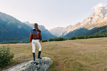 Obraz premium Woman stands on rock overlooking mountains landscape and nature meadow, hiking outdoors enjoying panoramic view at sunrise. Traveler in sweater and boots exploring alpine valley.