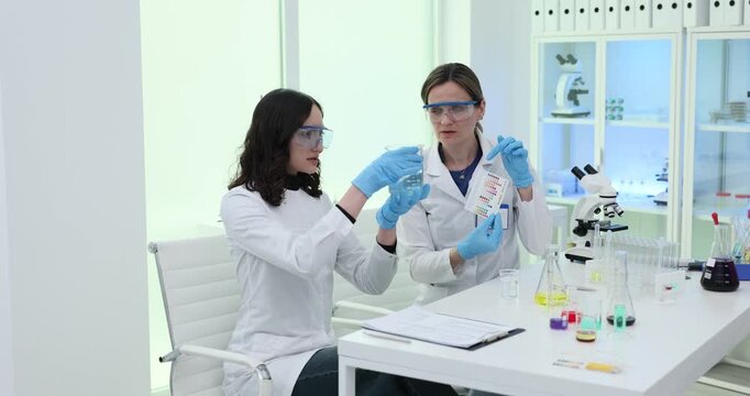 Female chemists analyze water properties using pH level test color charts in lab. Assistants examine liquid quality by modern equipment in laboratory