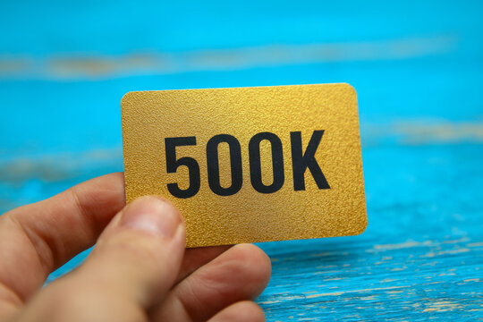 Hand holding a small golden card with bold black 500k text against a vibrant, textured blue wooden background symbolizing milestone achievement or value