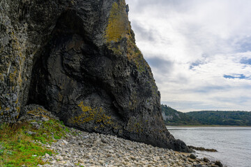 Beneath a dramatic sky, large rugged cliffs tower over smooth stones lining the tranquil shore of Russia's coastline. The calm waters reflect the surrounding greenery and clouds.