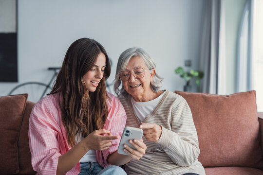 A young woman embraces her elderly mother as they sit on the sofa at home, smiling and looking at a phone together while watching content or shopping online in a relaxed family moment
