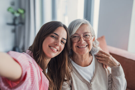 Smiling middle aged mother and daughter taking selfie together, hugging and sitting on couch at home, young woman and happy older mature mum photographing, two generations good relations