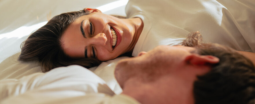 Beautiful happy loving young smiling couple relaxing in bed, looking at each other. Cozy home atmosphere, tenderness, closeness. Embracing, kissing. Lazy weekend, slow living concept