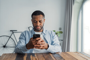 Confused African guy holding smart phone feels concerned thinking over received message. Mobile...
