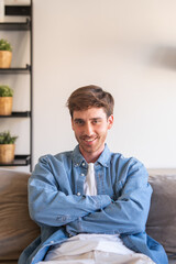 Naklejka premium Young caucasian man indoor sitting on couch smiling and looking at the camera with confidence, crossed arms enjoying the moment. Cheerful guy at home having fun