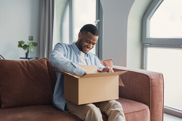 Smiling African American cheerful man unpacking parcel, sitting on couch at home, happy satisfied...