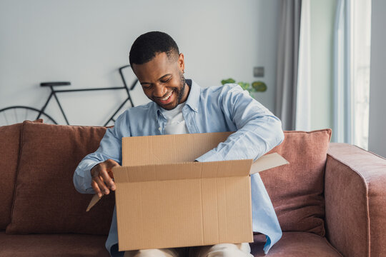 A smiling African American man sits on the couch at home while unpacking a parcel, looking happily inside a cardboard box after receiving an online order with a satisfying delivery experience