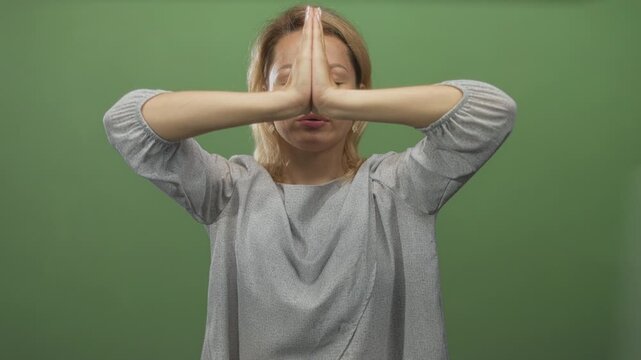 Young blonde woman in loose grey top presses hands in prayer in studio against green backdrop; serenity.