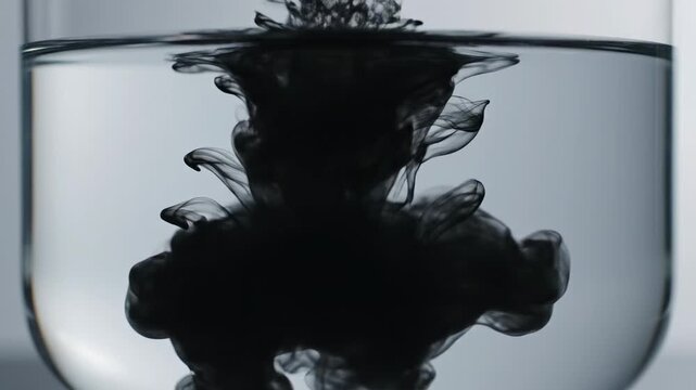 Ink dispersing in water. Black ink released into clear water inside a transparent glass container, spreading gradually in organic shapes.