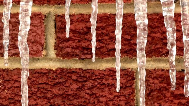 Closeup of clear icicles hanging from a house gutter against a red brick wall, showing winter ice dam formation and cold weather conditions.