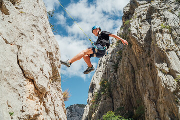 Climber teenage boy in protective helmet jumping on vertical cliff rock wall using rope Belay device, climbing harness in Paklenica canyon in Croatia. Active extreme sports time spending concept