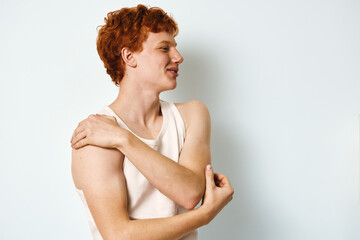 Obraz premium Young man with red hair in white tank top standing against white background, touching shoulder and looking sideways with relaxed and thoughtful expression.