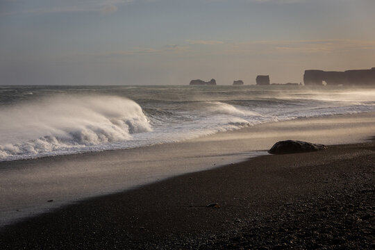 Sneaker waves crushing against Reynisfjara Black Sand Beach coast, with strong winds blowing water and volcanic black sand, sunset, extreme weather, Iceland.