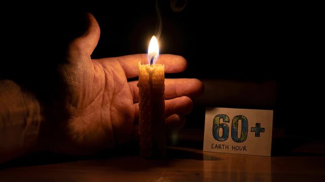 Mans hand holding lit candle beside earth hour sign. Environmental conservation symbol for 60 plus awareness event. Eco friendly lighting and energy saving concept in dark setting.