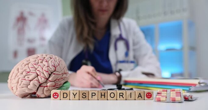Wooden cubes spell word Dysphoria near brain model. Female psychiatrist writes report while tracking mood disorder symptoms during patient visit