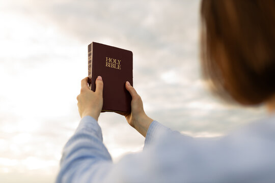 Hands raised holding a Bible under a cloudy sky